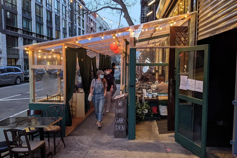 Person wearing mask walking through temporary outdoor dining setup lined with bistro lights at dusk in New York City.